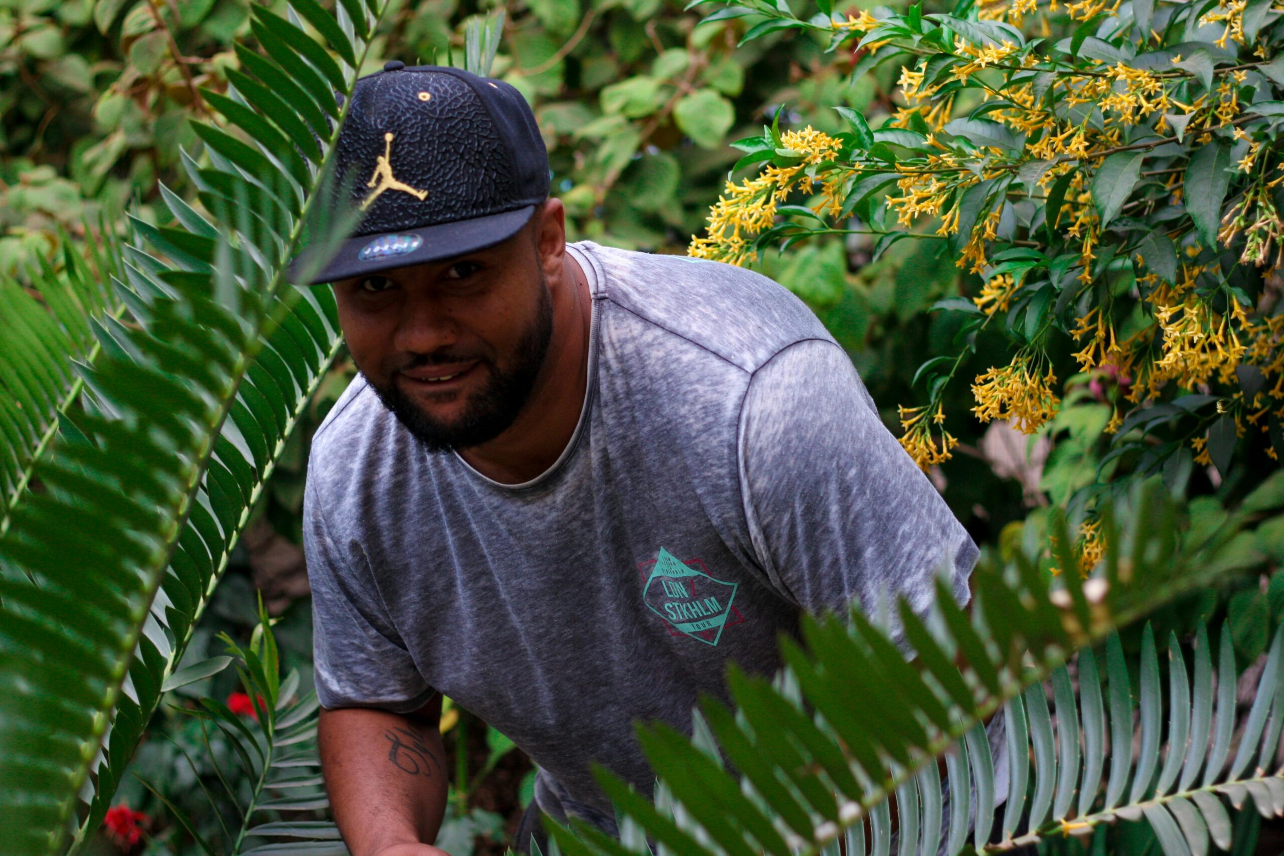 A man wearing a baseball cap smiles in a lush garden with vibrant flora.